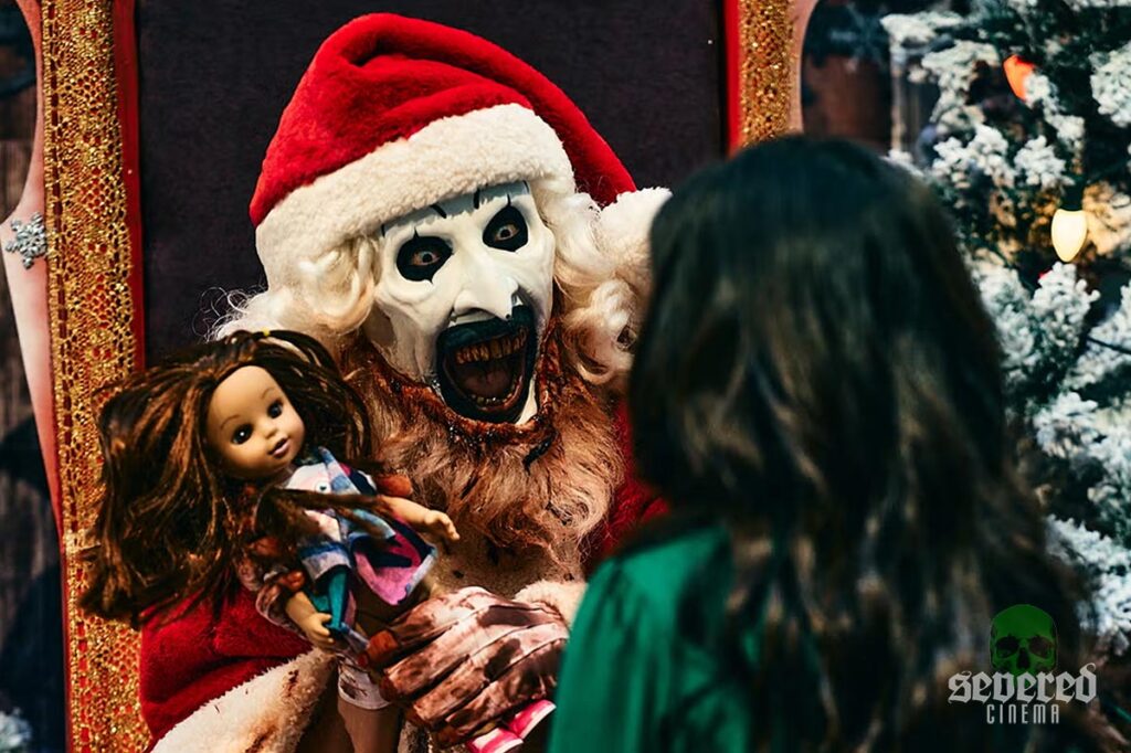 Art the Clown dressed as Santa holding a doll for a little girl from Terrifier 3