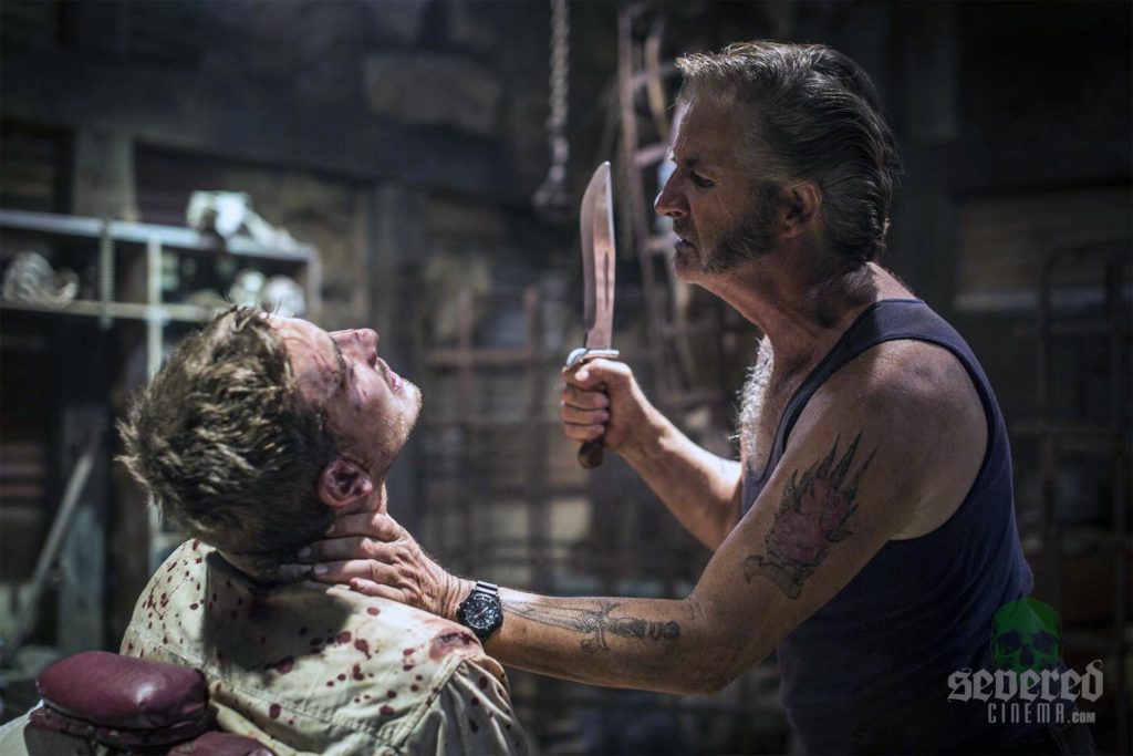 Photo of John Jarret as Mick Taylor in Wolf Creek 3 holding a knife and grabbing a persons throat.